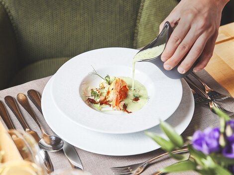 Ein elegantes Gericht wird auf einem weißen Teller serviert. Eine Hand gießt eine grüne Sauce über das Essen, das mit Kräutern garniert ist.