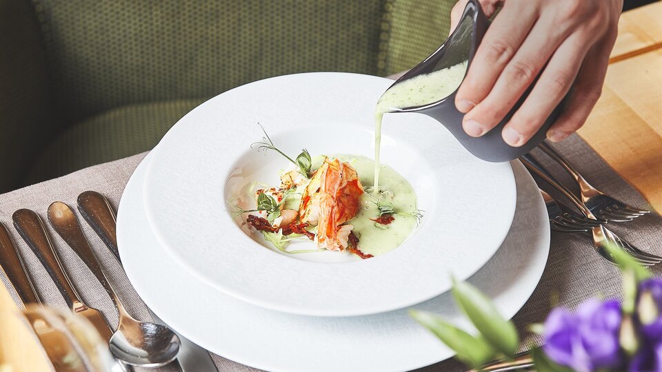 Ein elegantes Gericht wird auf einem weißen Teller serviert. Eine Hand gießt eine grüne Sauce über das Essen, das mit Kräutern garniert ist.