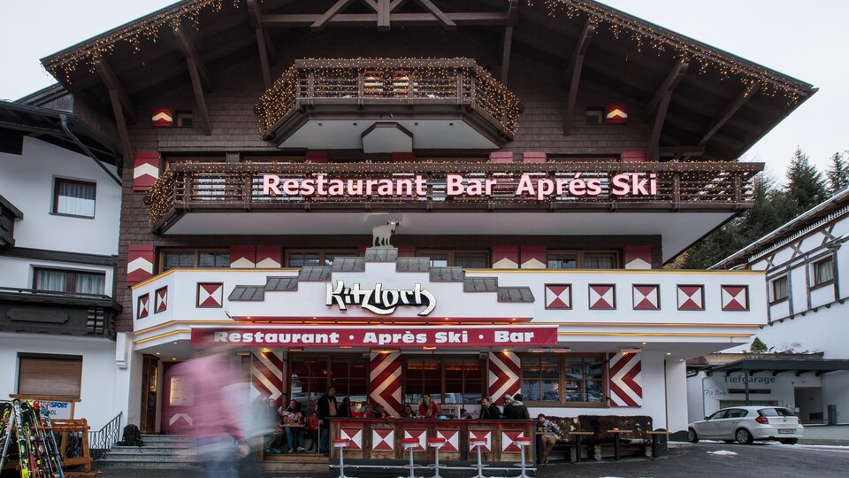 Ein gemütliches Restaurant mit einer Bar im Après-Ski-Stil. Die Fassade ist rustikal mit Holzdetails und einem einladenden Außenbereich.