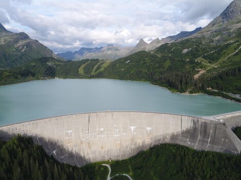 An impressive dam surrounded by green mountains and a clear lake. The scenery depicts a tranquil natural backdrop with a slightly cloudy sky.