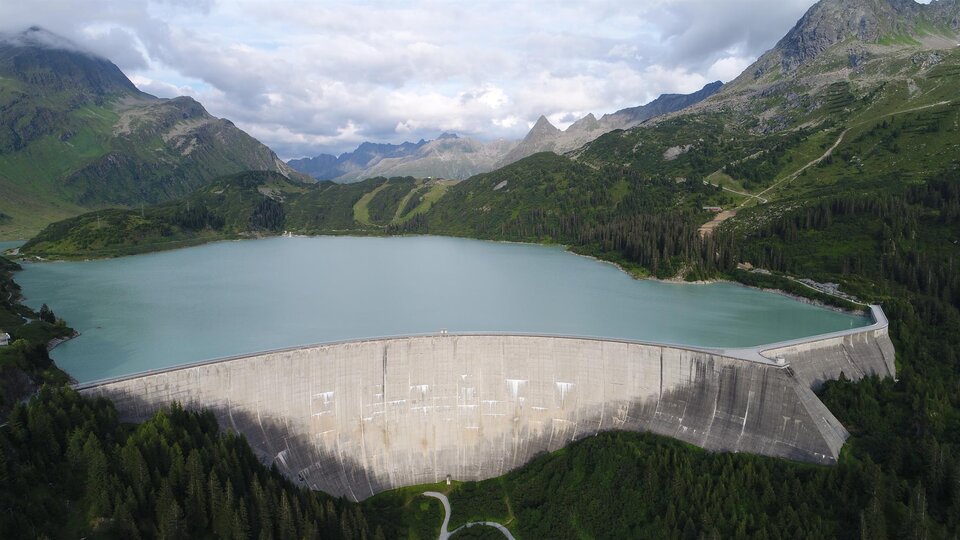 Ein beeindruckender Staudamm umgeben von grünen Bergen und einem klaren See. Die Szenerie zeigt eine ruhige Naturkulisse mit leicht bewölktem Himmel.