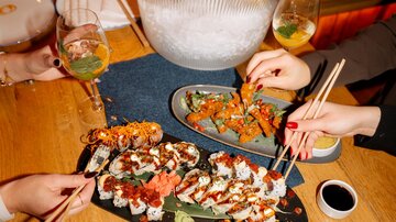 Ein Tisch mit Sushi-Platten und Gläsern mit Getränken. Im Hintergrund steht eine Eisbox mit Sekt.