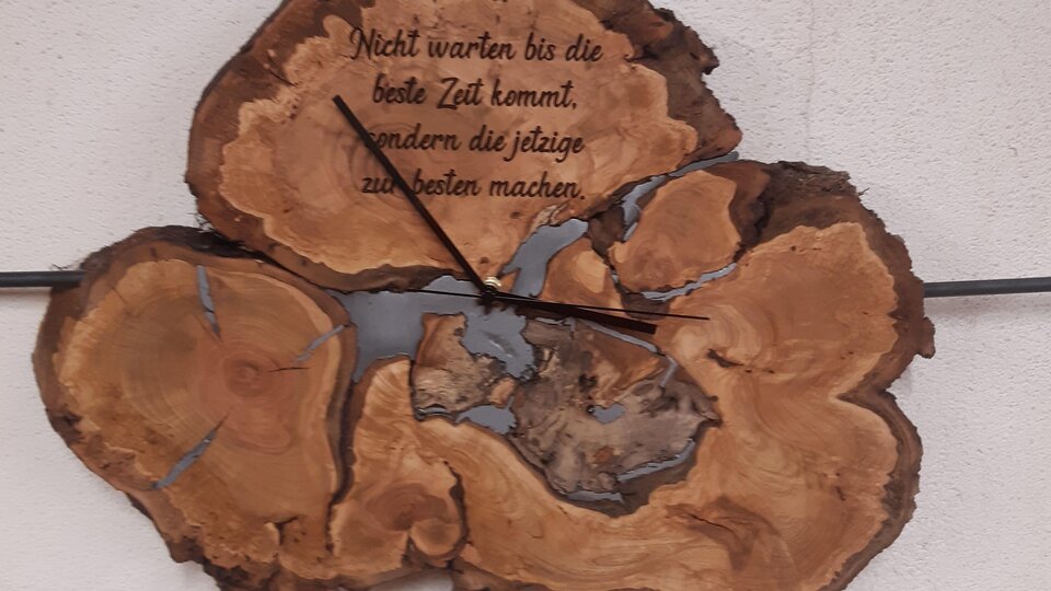 Eine dekorative Wanduhr aus Holz mit einer einzigartigen Form. Darauf steht der Spruch: "Nicht warten bis die beste Zeit kommt, sondern die jetzige zu nutzen machen."