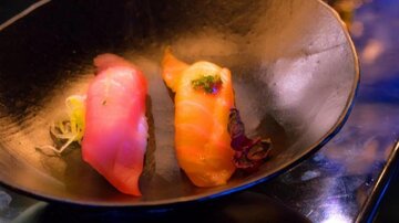 Zwei Sushi-Stücke auf einem schwarzen Teller. Das eine Stück ist rosa und das andere orange, garniert mit Kräutern.