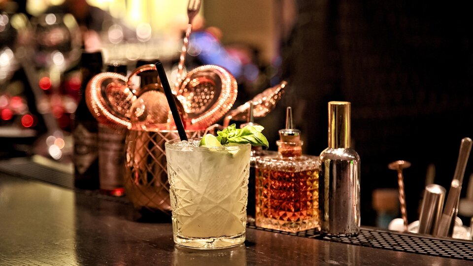 Ein leckerer Cocktail mit frischer Minze steht auf einer eleganten Bar. Im Hintergrund sind verschiedene Flaschen und Utensilien zu sehen.