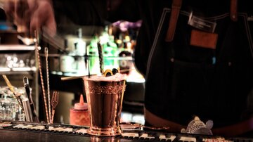 Ein Barkeeper bereitet einen Cocktail in einer stilvollen Bar vor. Im Hintergrund sind verschiedene Getränke in leuchtenden Farben zu sehen.