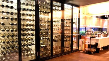 Ein stilvoller Weinkeller mit großen Glasregalen, die eine Vielzahl von Weinen präsentieren. Im Hintergrund ist eine Bar mit verschiedenen Getränken zu sehen.
