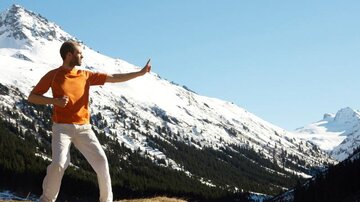 Ein Mann in orangefarbenem Shirt steht auf einer Wiese in den Bergen. Im Hintergrund sind schneebedeckte Gipfel und ein klarer blauer Himmel zu sehen.
