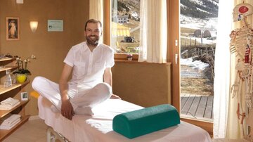 Ein freundlicher Therapeut sitzt auf einer Massageliege in einem hellen Raum. Im Hintergrund sieht man eine schöne Berglandschaft durch das Fenster.