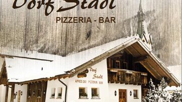 Ein gemütliches Gebäude im alpinen Stil mit dem Namen "Dorf Stadl". Es handelt sich um eine Pizzeria und Bar, umgeben von schneebedeckten Bergen.
