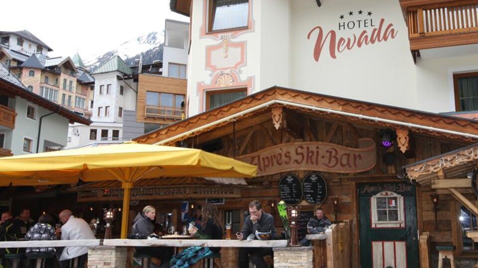 Ein gemütliches Restaurant mit einer Holzterrasse und einem großen Sonnenschirm. Menschen sitzen zusammen und genießen Getränke, während die Berge im Hintergrund sichtbar sind.