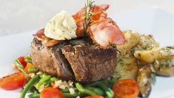 Ein saftiges Steak auf einem Bett aus Gemüse und Kartoffeln, garniert mit Speck und Kräuterbutter. Frische Tomaten und grüne Bohnen runden das Gericht ab.