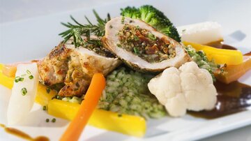 Ein kunstvoll angerichteter Teller mit gefülltem Hähnchen, umgeben von buntem Gemüse wie Karotten, Brokkoli und Blumenkohl. Auf dem Teller liegt auch grüne Risotto und eine schmackhafte Sauce.