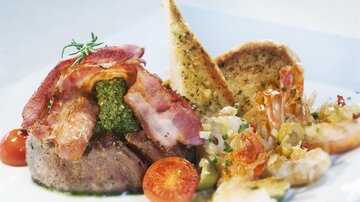 Ein zartes Steak, umhüllt von knusprigem Speck und serviert mit Garnelen, Tomaten und geröstetem Brot. Das Gericht ist stilvoll angerichtet und sieht appetitlich aus.
