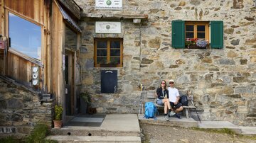 Eine gemütliche Berghütte aus Stein mit grünen Fensterläden. Ein Paar sitzt entspannt vor der Hütte mit einer Tasche daneben.