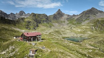 Eine malerische Berglandschaft mit einer Berghütte und einem klaren See. Die Umgebung ist grün und von beeindruckenden Bergen umgeben.