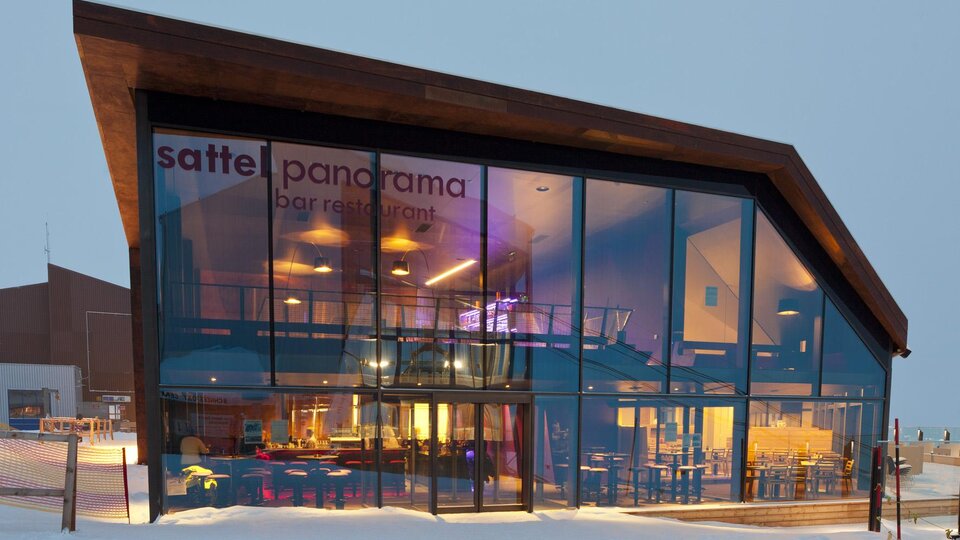 Ein modernes Restaurant mit großen Glasfenstern und einem schlichten Holzdesign. Das Gebäude liegt in einer winterlichen Umgebung mit Schnee.