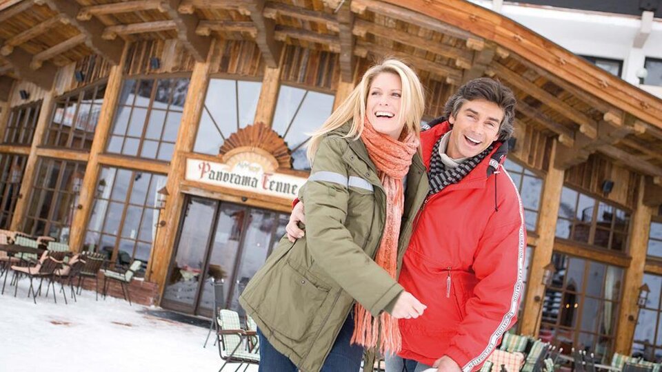 Ein glückliches Paar steht lächelnd vor einem Holzgebäude im Schnee. Im Hintergrund ist eine Terrasse mit Stühlen und Tischen zu sehen.