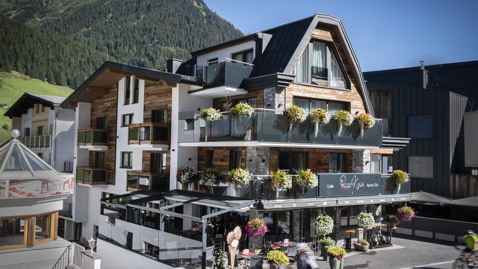 Ein modernes Hotel mit blühenden Balkonpflanzen in einer Berglandschaft. Die Fassade ist freundlich und einladend.