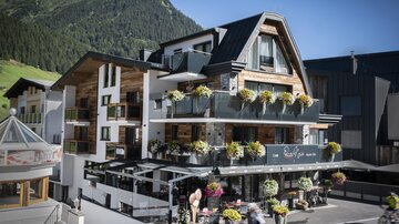 Ein modernes Hotel mit blühenden Balkonpflanzen in einer Berglandschaft. Die Fassade ist freundlich und einladend.