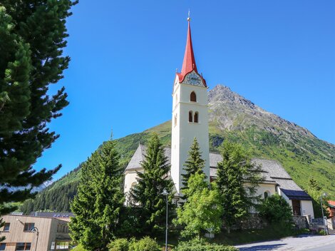 A picturesque church with a red tower, surrounded by green trees and mountains. The sky is clear and blue. | © TVB Paznaun - Ischgl