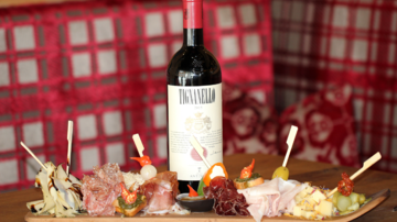 Eine Flasche Rotwein steht auf einem Tisch, umgeben von einer Auswahl an Wurst- und Käseplatten. Der Hintergrund zeigt eine gemütliche, rustikale Holzvertäfelung.