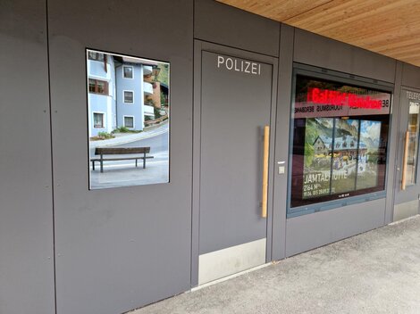 A modern police station entrance with a large window. Next to the entrance is a picture of a bank and residential buildings.