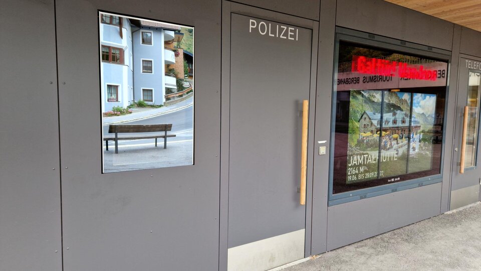 Ein moderner Polizeigebäudeeingang mit einem großen Fenster. Neben dem Eingang ist ein Bild einer Bank und Wohngebäude zu sehen.