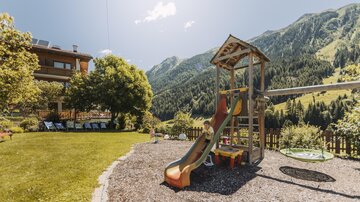 Ein Spielplatz mit einer Rutsche und Schaukeln in einer grünen Wiese. Im Hintergrund sind Berge und ein blauer Himmel zu sehen.
