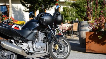 Ein schwarzes Motorrad steht vor einem gemütlichen Restaurant mit Blumen und Tischen im Freien. Im Hintergrund sind grüne Bäume und ein schön gestalteter Außenbereich sichtbar.