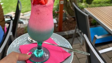 Ein bunten Cocktail mit Erdbeeren und einer Waffel in einem gemütlichen Außenbereich. Im Hintergrund ist eine grüne Gartenlandschaft sichtbar.