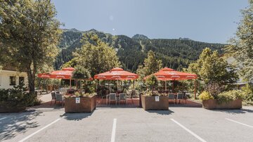 Ein einladendes Café mit roten Sonnenschirmen, umgeben von Bäumen. Im Hintergrund sind schattige Berge zu sehen.