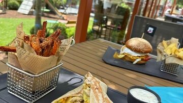 Ein Tisch im Freien mit verschiedenen Gerichten. Zu sehen sind ein Wrap, Pommes, ein Burger und knusprige Chicken Wings in einer Fritteuse.