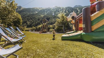 Ein schöner Spielplatz im Freien mit einem großen Hüpfburg und einer malerischen Berglandschaft im Hintergrund. Im Vordergrund stehen Liegestühle auf dem Gras.