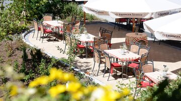 Ein einladendes Restaurant im Freien mit Tischen und Stühlen unter weißen Sonnenschirmen. Die Umgebung ist grün und blühend, ideal zum Entspannen und Genießen.