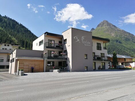 A modern building in the mountains with a clear blue sky. The house contains several apartments and is surrounded by green forests.