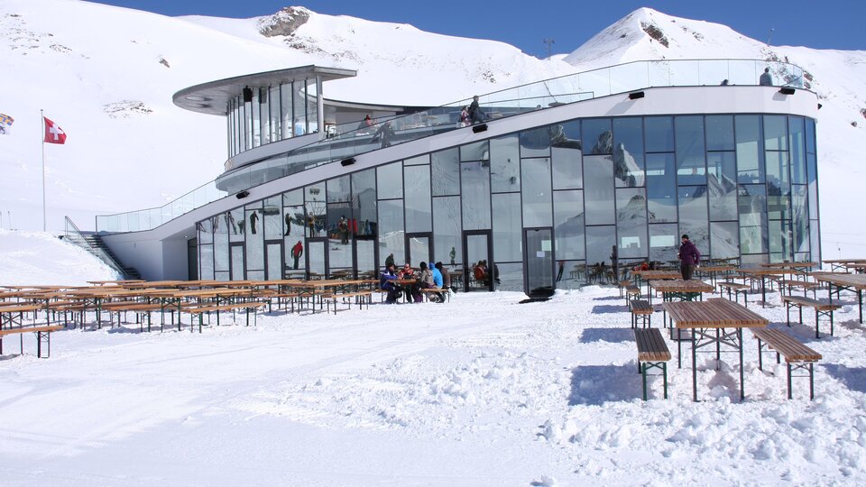 Ein modernes Gebäude in den Bergen, umgeben von Schnee. Draußen stehen Holzbankreihen und Menschen genießen die winterliche Landschaft.