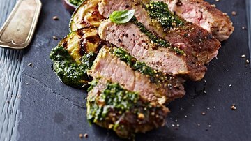 Ein köstliches Stück Fleisch, perfekt zubereitet und mit grünem Pesto garniert. Dazu liegen gegrillte Zucchini auf einem dunklen Servierbrett.
