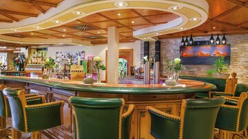 Eine elegante Bar mit einer geschwungenen Theke und gemütlichen grünen Barhockern. Die warme Holzdecke und die dekorativen Pflanzen schaffen eine einladende Atmosphäre.
