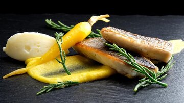 Ein kunstvoll angerichteter Teller mit gegrilltem Fisch, umgeben von buntem Püree und frischem Gemüse. Die Präsentation nutzt elegante Elemente wie Kräuter und eine dunkle Unterlage für einen modernen Look.