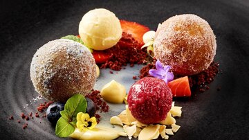 Ein kunstvoll angerichteter Dessertteller mit drei gefüllten Kugeln in verschiedenen Geschmacksrichtungen. Umgeben von bunten Früchten und dekorativen Elementen.