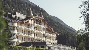 Ein elegantes Hotel mit einem schönen Balkon und spitzen Dächern liegt malerisch in den Bergen. Die umgebende Landschaft ist grün und bewaldet.
