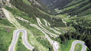 A winding road leads through a green mountain landscape. The mountains and the valley are covered with trees and the sky is clear.