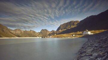 Eine ruhige Seenlandschaft umgeben von hohen Bergen und dramatischen Wolkenformationen. Im Vordergrund ist eine steinige Uferlinie mit einem weißen Gebäude sichtbar.