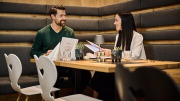 Ein gemütliches Café mit einem holzgetäfelten Ambiente. Zwei Personen sitzen an einem Tisch und unterhalten sich, während sie die Speisekarten ansehen. | © Silvrettaseilbahn AG
