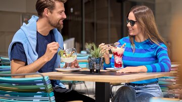 Ein Paar sitzt an einem Tisch im Freien und genießt Desserts und Cocktails. Die Szene ist hell und einladend, mit fröhlichem Wetter.
