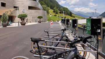Eine moderne Fahrradstation mit E-Bikes steht vor einem eleganten Gebäude. Im Hintergrund sind grüne Hügel und ein blauer Himmel zu sehen.