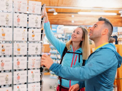 A couple is looking for souvenirs in a store. The woman reaches for an item while the man watches with interest. | © Golm Silvretta Lünersee Tourismus GmbH Bregenz, Christoph Schoech