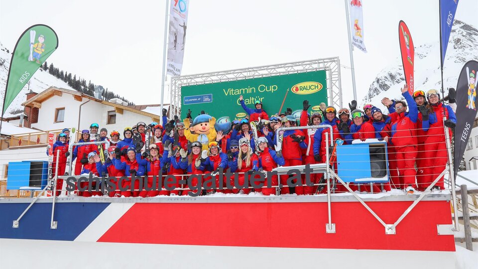 Eine Gruppe von Skifahrern in bunten Anzügen steht auf einer Tribüne. Im Hintergrund sind Schnee und Fahnen zu sehen, die für den Vitamin Cup werben.
