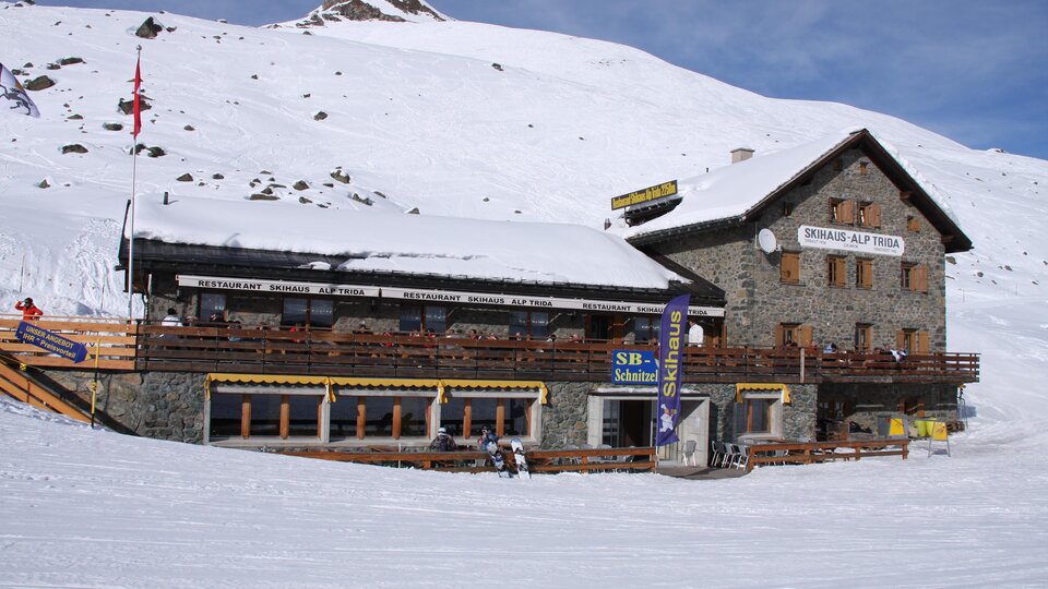 Ein gemütliches Berghaus im Schnee, umgeben von einer winterlichen Landschaft. Die Fassade ist aus Stein mit einem großen Balkon und sonnigen Terrassen.